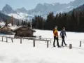 Coppia cammina in una località di montagna trentino inverno, tra baite di legno e montagne innevate.