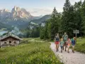 Famiglia in un percorso di trekking facile tra prati fioriti e montagne maestose, con un rifugio alpino e mucche al pascolo.