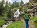 Un escursionista ammira una cascata nel Val di Sole in estate, con un ponte di legno che attraversa il torrente.