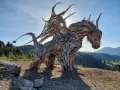 Un maestoso drago di Vaia, scolpito nel legno, domina un paesaggio montano con le ali spiegate verso il sole.