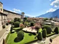 Giardini curati e fioriti incorniciano il maestoso Castel Buonconsiglio, con torri imponenti e montagne sullo sfondo.