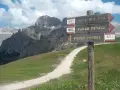Cartello con indicazioni per Viel del Pan, Passo Fedaia e Passo Pordoi. Sentiero in salita tra prati verdi e montagne imponenti.