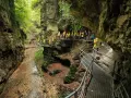 Un gruppo di turisti con impermeabili gialli esplora il **rio sass fondo**, un canyon verdeggiante con passerelle metalliche che si snodano tra le rocce.