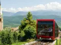 Funicolare rossa in Trentino Alto Adige, con campanile e montagne sullo sfondo. Un'ottima idea per scoprire cosa vedere!