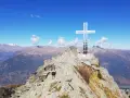 Escursionista raggiunge la vetta con la croce della pace, circondata da montagne maestose sotto un cielo azzurro.