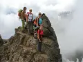Quattro escursionisti con zaini si fermano sulla cima rocciosa di Cima Sternai, circondati da nuvole e nebbia.