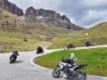 Motociclisti affrontano tornanti sui **Dolomiti passi**, con imponenti vette sullo sfondo.