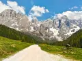 Sentiero sterrato tra prati verdi e boschi, con imponenti vette innevate sullo sfondo. Le Dolomiti facili invitano a una passeggiata panoramica.