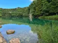 Acque cristalline del lago di Lavis riflettono la rigogliosa vegetazione. Rocce affiorano, un corridore si allena lungo la riva.