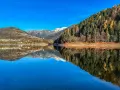 Il lago delle Piazze riflette montagne innevate, boschi autunnali e un villaggio sotto un cielo azzurro.