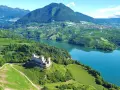 Castello medievale su una collina verdeggiante, con vigneti a terrazze e un lago azzurro nel **Val di Non**.