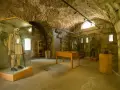 Interno di un museo in pietra con manichini in uniformi storiche, uno dei quali indossa un abito chiaro e porta un fucile, evocando un forte strino di battaglia.
