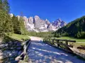 Ponte in legno su ruscello, sentiero per trekking Val Venegia, con montagne innevate e boschi verdi sotto un cielo azzurro.