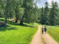 Due persone camminano su un sentiero nella Val di Non, circondate da prati verdi e alberi. Una baita di legno si intravede tra gli alberi.