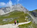 Escursionisti su sentiero ghiaioso, godendosi un **trekking più giorni Dolomiti** con vette imponenti e prati verdi.