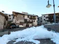 Paesaggio innevato a Miola di Pinè, con case in pietra e legno, auto parcheggiate e un cielo azzurro.