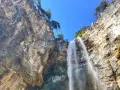 La cascata di Tret scende maestosa da una parete rocciosa imponente, sotto un cielo azzurro e limpido.