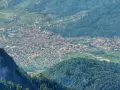 Vista aerea della Valsugana, Trento, con un paese incastonato tra montagne verdi e un picco roccioso in primo piano.