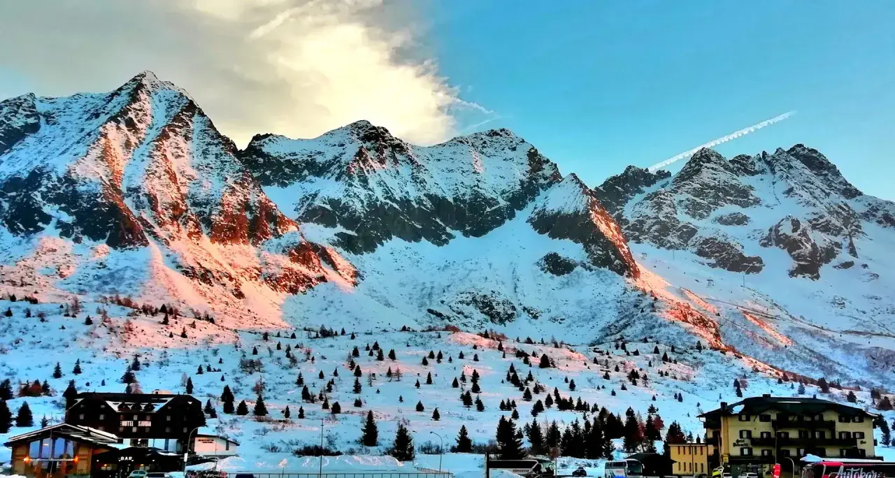 Montagne innevate al tramonto sul Passo del Tonale, con alberi sparsi e un hotel.