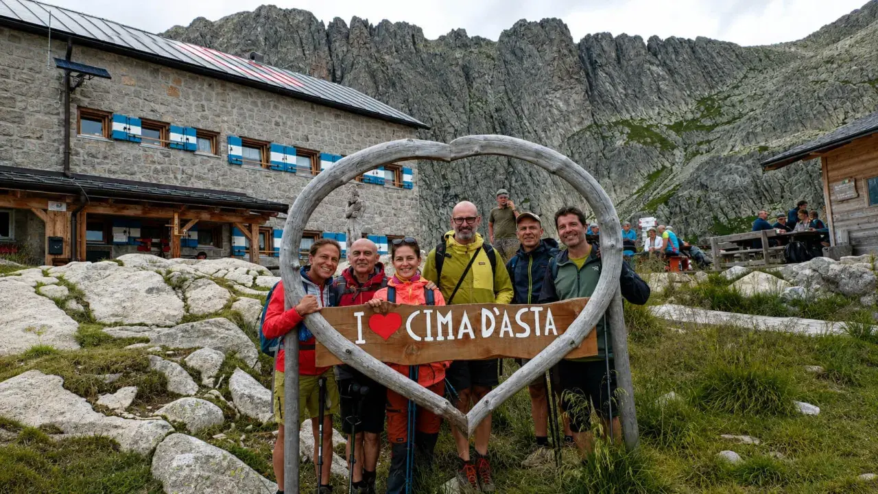 Gruppo di escursionisti felici davanti a un rifugio alpino, con un cartello a forma di cuore che dice "I ❤️ CIMA D'ASTA". Un'indimenticabile esperienza di trekking Trentino.