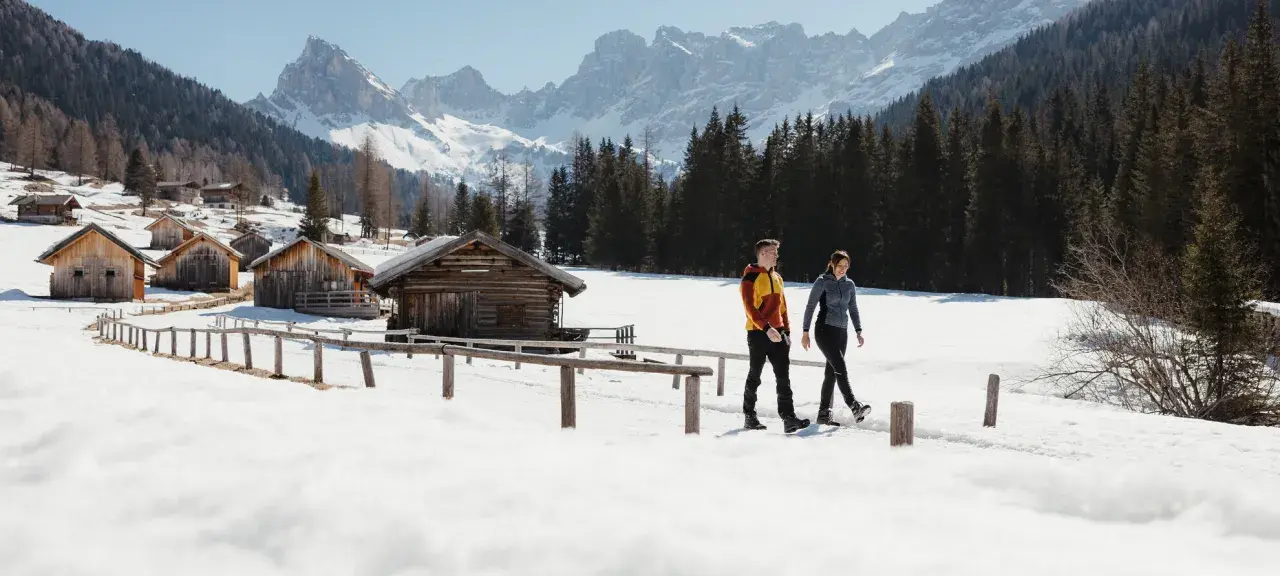 Coppia cammina in una località di montagna trentino inverno, tra baite di legno e montagne innevate.