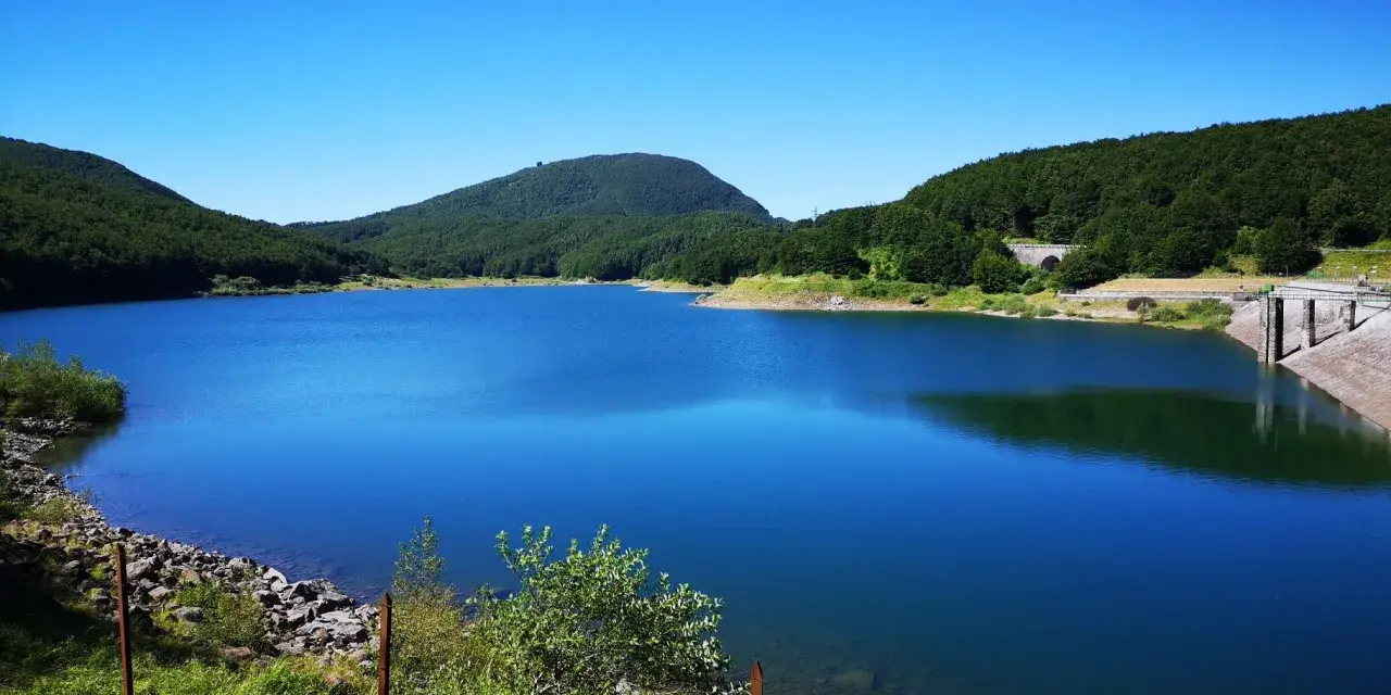 Vasto lago di Comano con acque blu intenso, circondato da colline verdi e una diga in cemento.