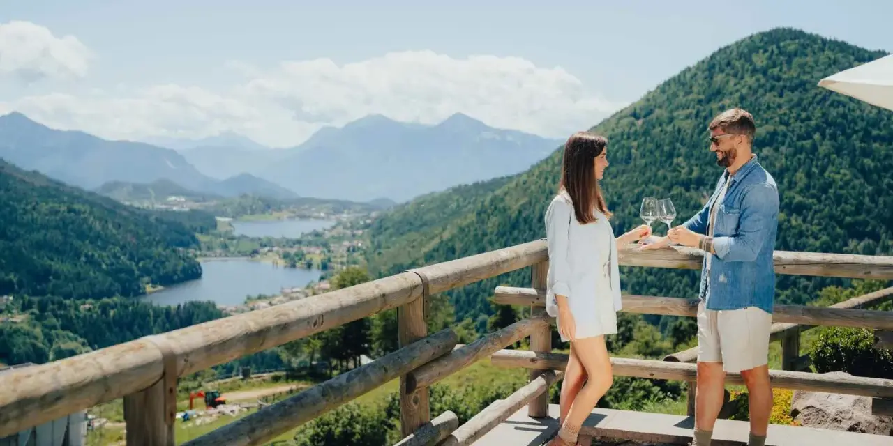 Coppia brinda con vino su un balcone con vista sulla **montagna di Pinè** e laghi.