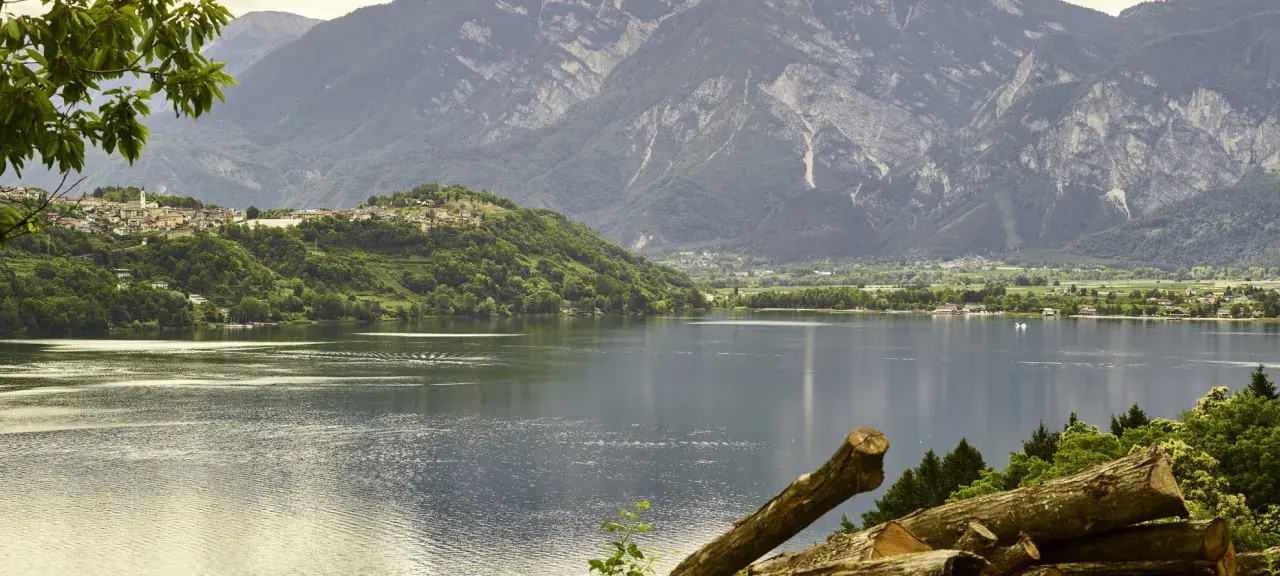 Villaggio su collina verde affacciato sul lago di Caldonazzo, con montagne imponenti sullo sfondo.