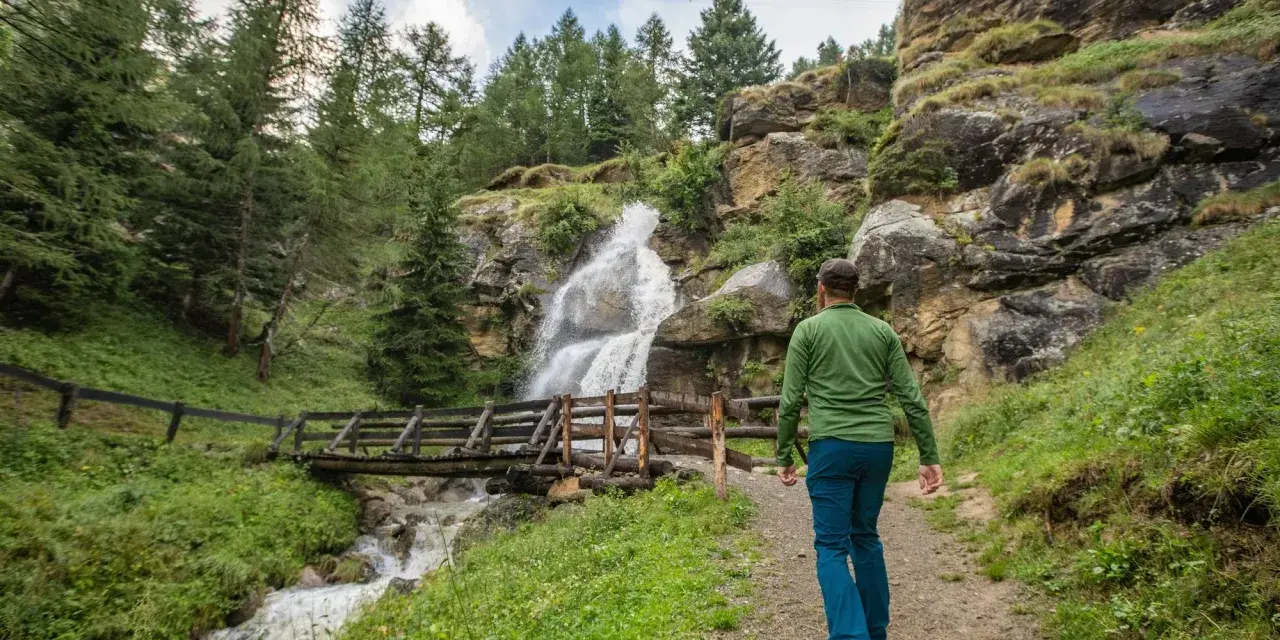 Un escursionista ammira una cascata nel Val di Sole in estate, con un ponte di legno che attraversa il torrente.