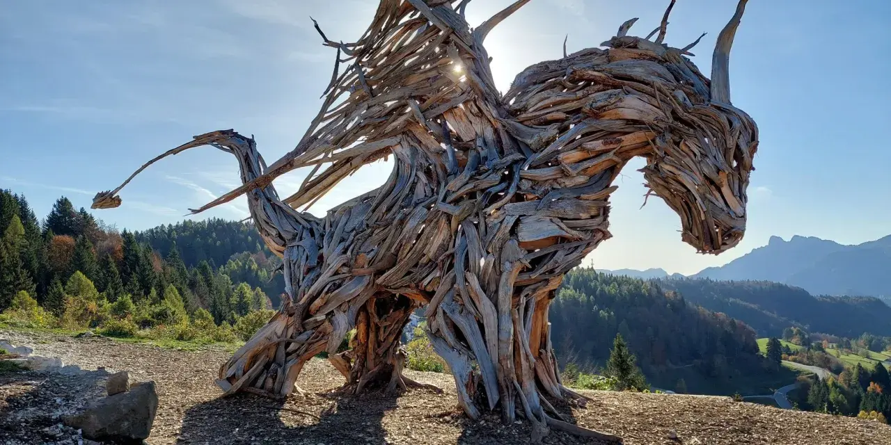 Un maestoso drago di Vaia, scolpito nel legno, domina un paesaggio montano con le ali spiegate verso il sole.