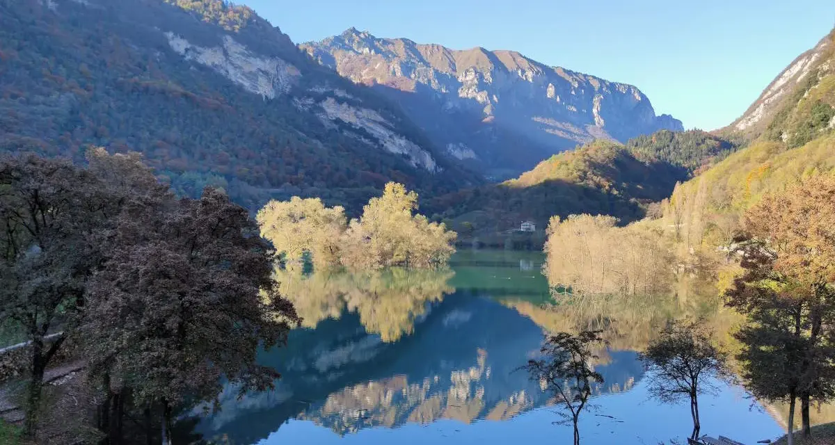 Il lago di Tenno riflette montagne imponenti e alberi autunnali. Due persone siedono su una panchina, godendosi la quiete.
