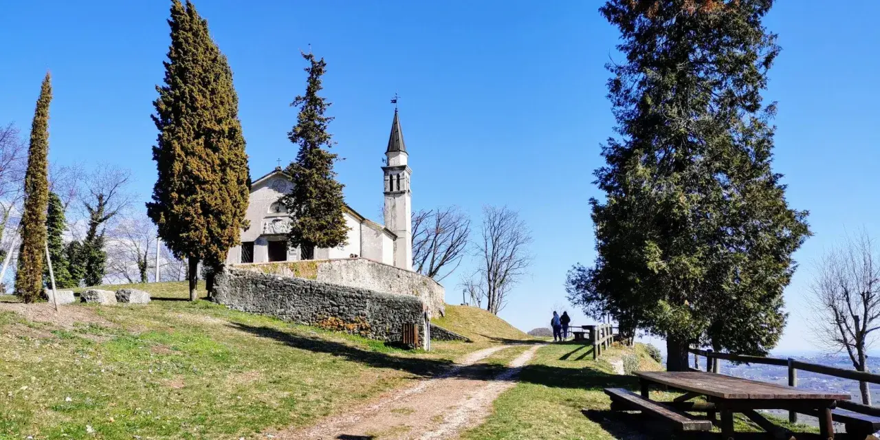 A church with a bell tower sits on a grassy hill, with tall trees and a picnic table in the foreground.