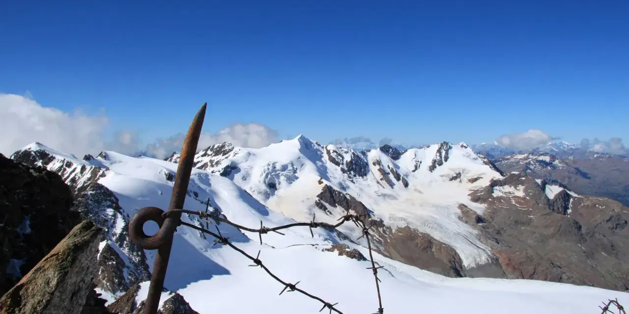 Vista mozzafiato dalla punta Linke, con cime innevate e un cielo azzurro intenso. Un vecchio piccone arrugginito e filo spinato segnano il confine.