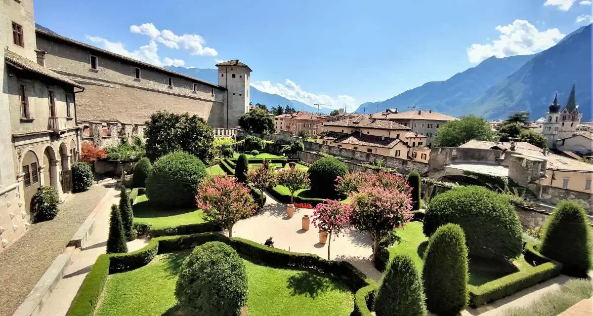 Giardini curati e fioriti incorniciano il maestoso Castel Buonconsiglio, con torri imponenti e montagne sullo sfondo.