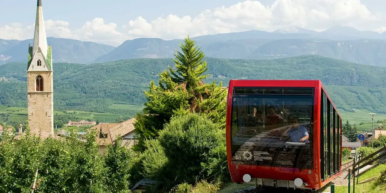 Funicolare rossa in Trentino Alto Adige, con campanile e montagne sullo sfondo. Un'ottima idea per scoprire cosa vedere!