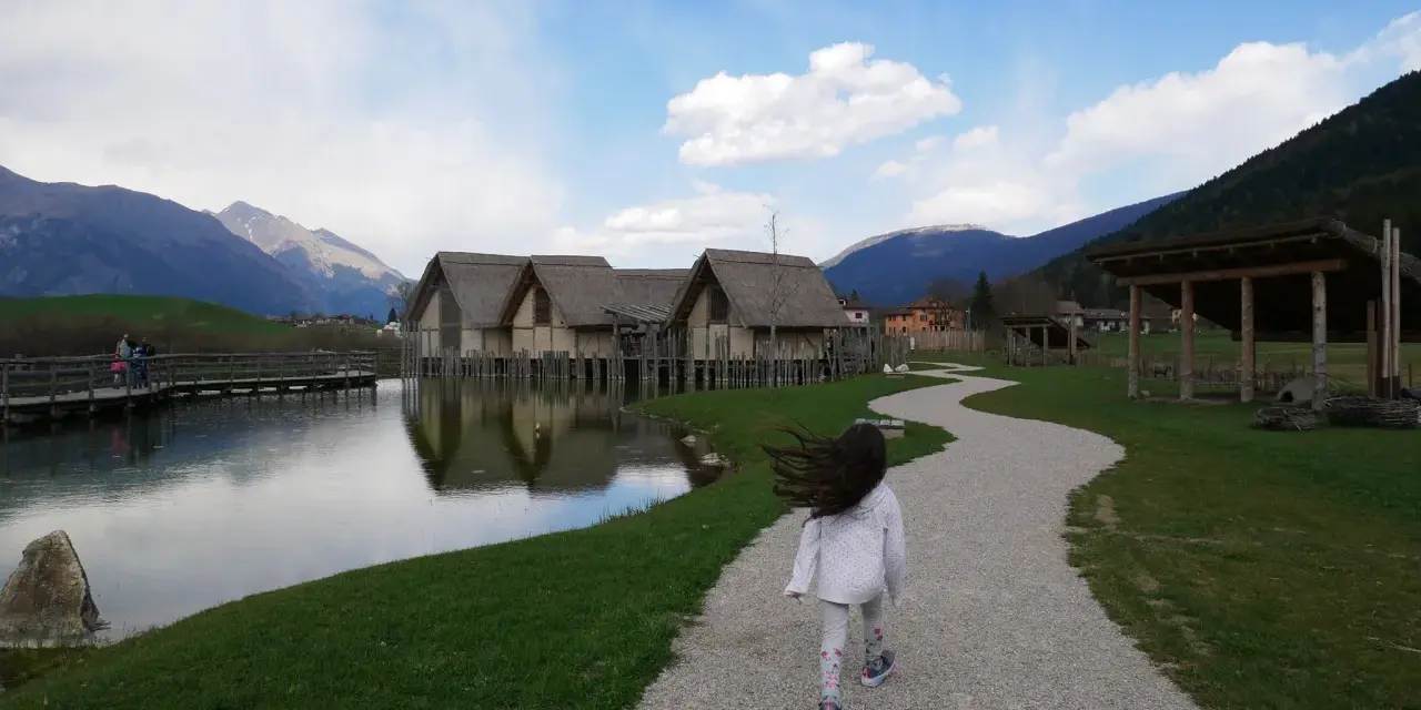Bambina corre su sentiero ghiaioso verso villaggio palafitticolo nelle Alpi Giudicarie. Montagne sullo sfondo.