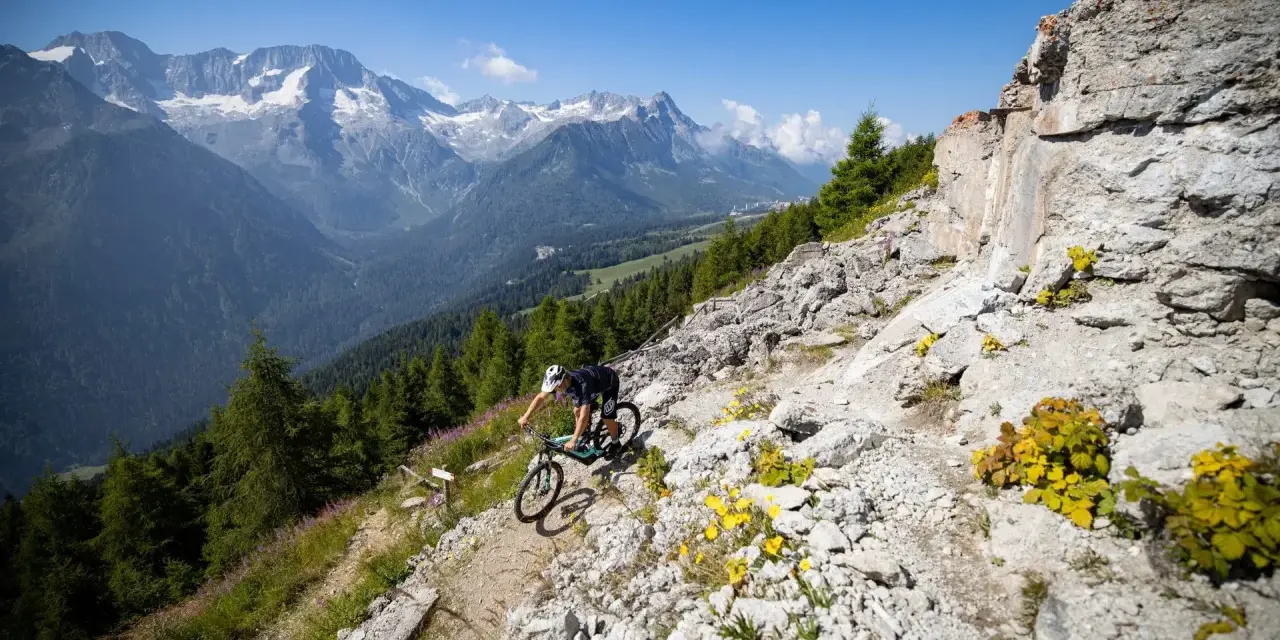 Ciclista in discesa su sentiero roccioso, con montagne innevate sullo sfondo, durante il giro dei forti tonale.