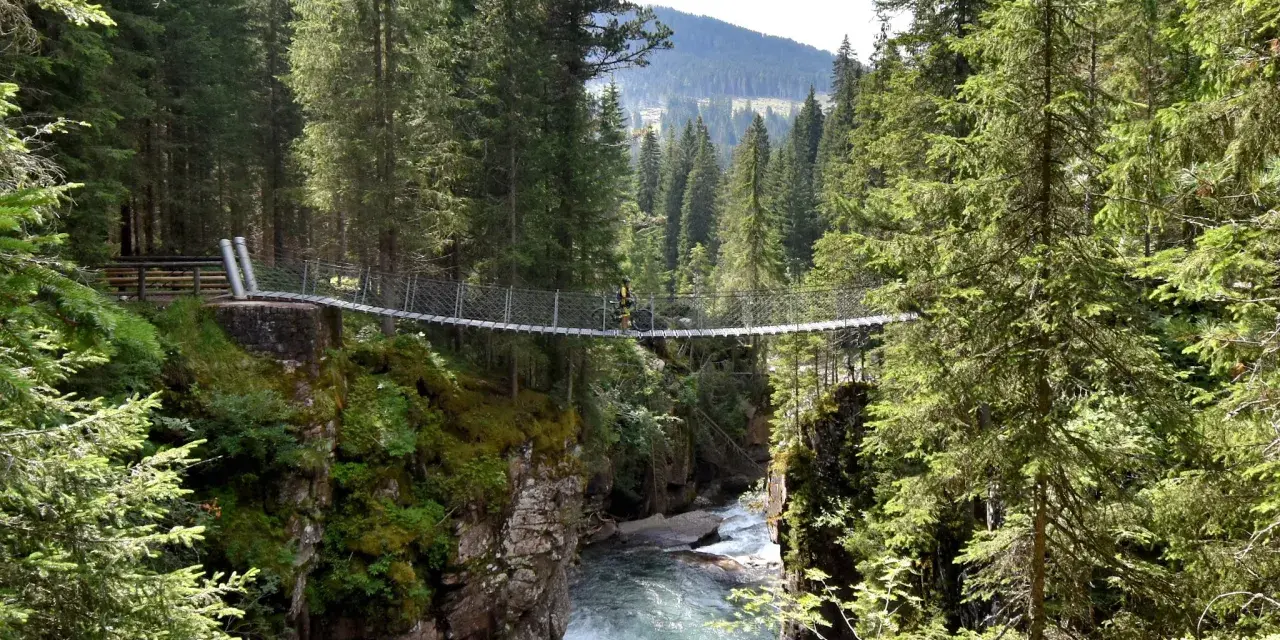Un ciclista attraversa un avventuroso ponte tibetano sospeso su un torrente impetuoso, circondato da una fitta foresta alpina.