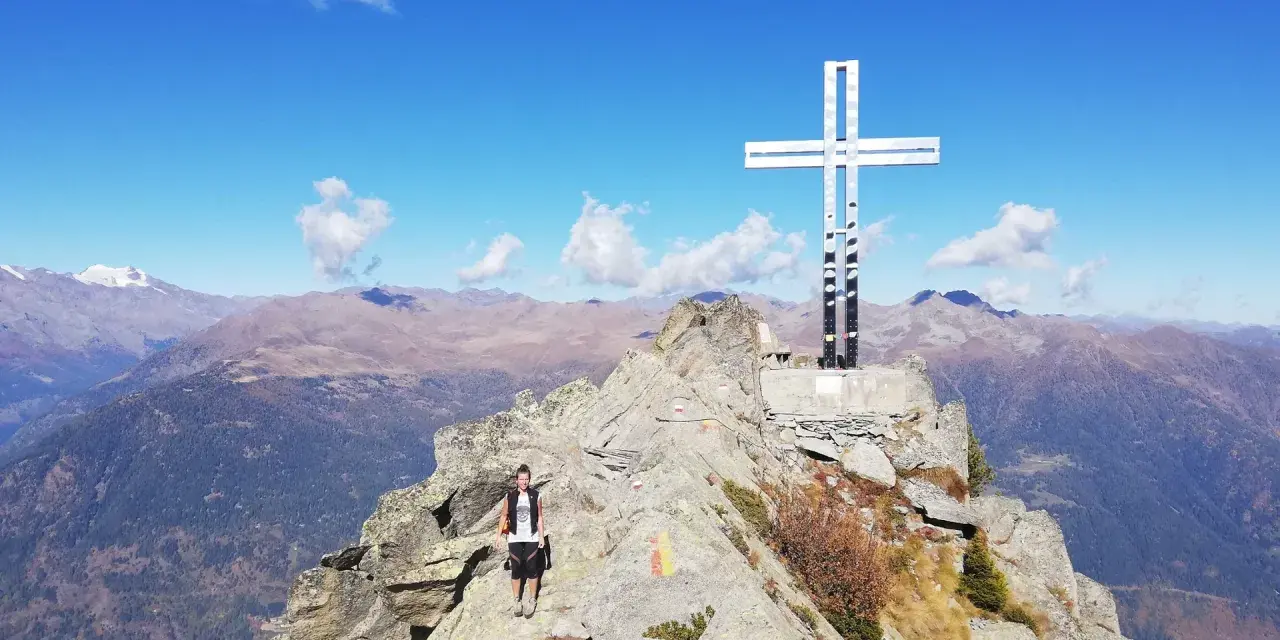 Escursionista raggiunge la vetta con la croce della pace, circondata da montagne maestose sotto un cielo azzurro.