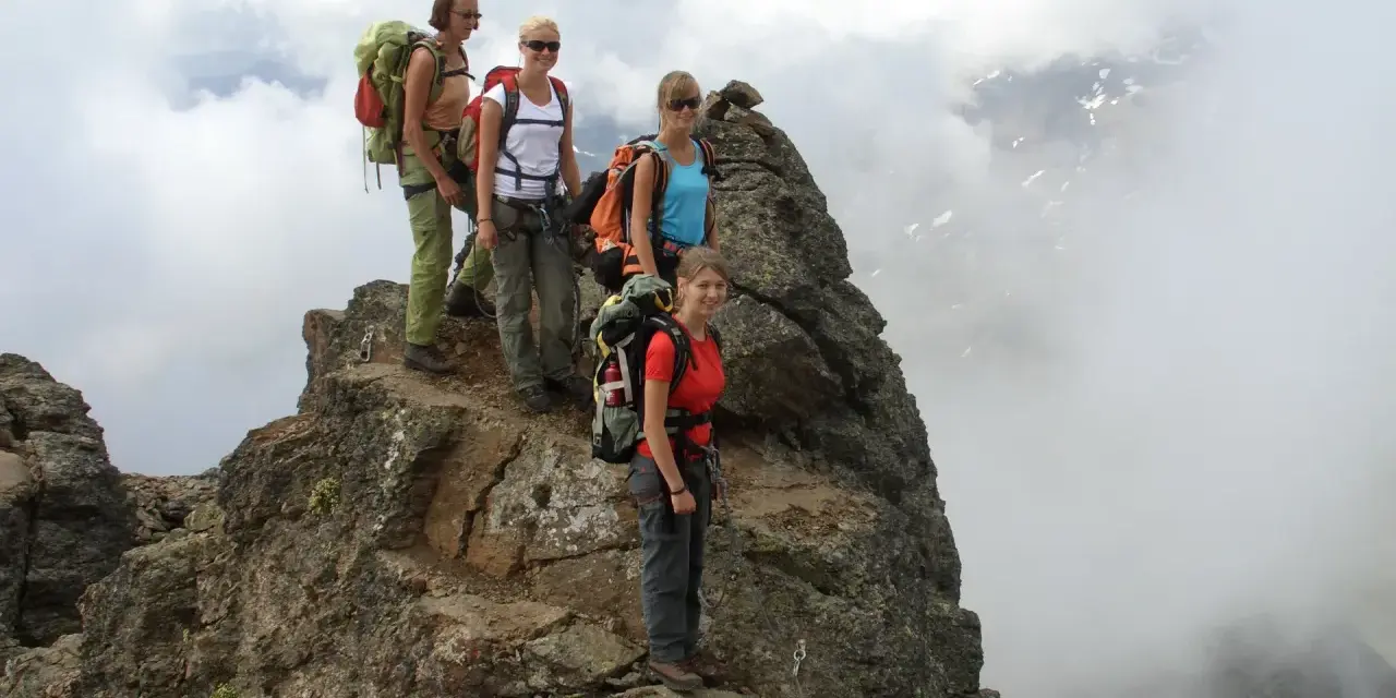 Quattro escursionisti con zaini si fermano sulla cima rocciosa di Cima Sternai, circondati da nuvole e nebbia.