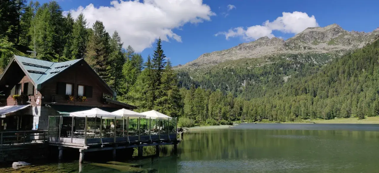 Rifugio in legno con tavoli all'aperto sul lago, circondato da boschi e montagne. Un luogo incantevole per scoprire cosa vedere a Pellizzano.