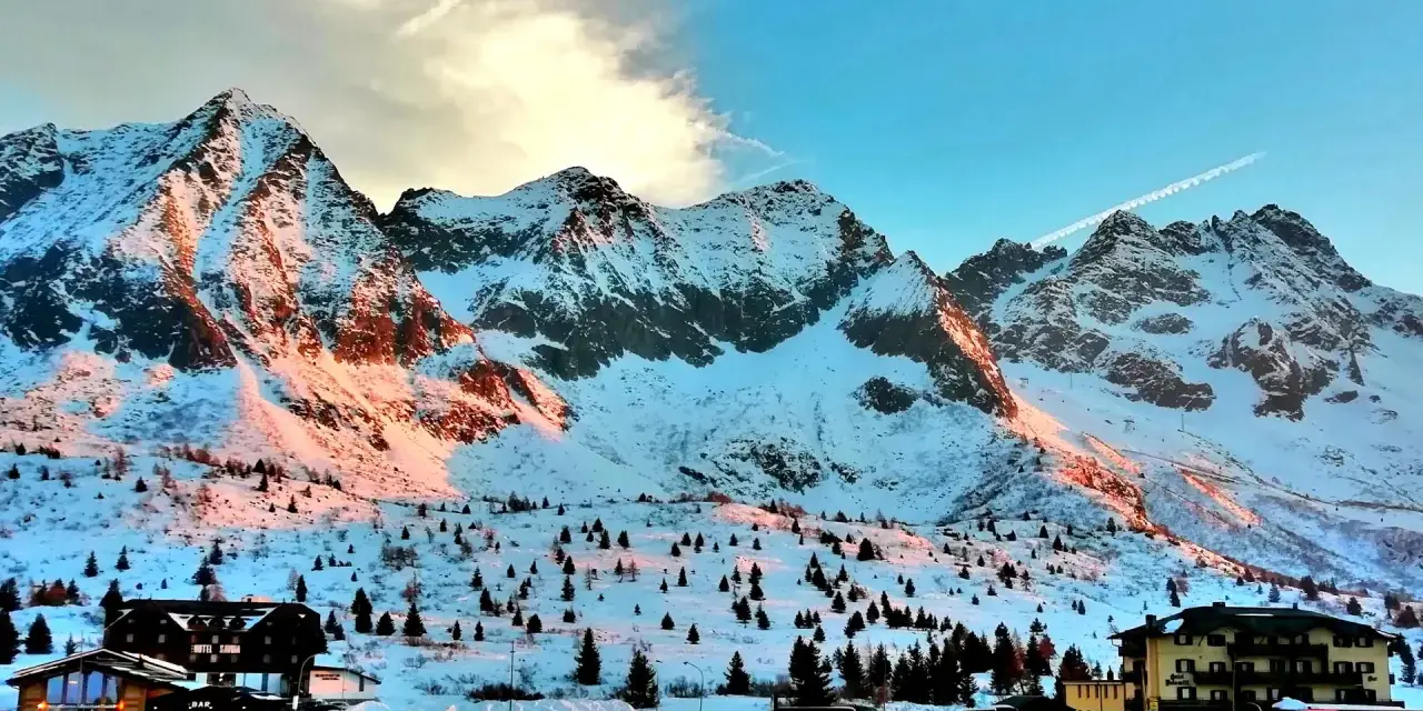 Montagne innevate al tramonto sul Passo del Tonale, con alberi sparsi e un hotel.