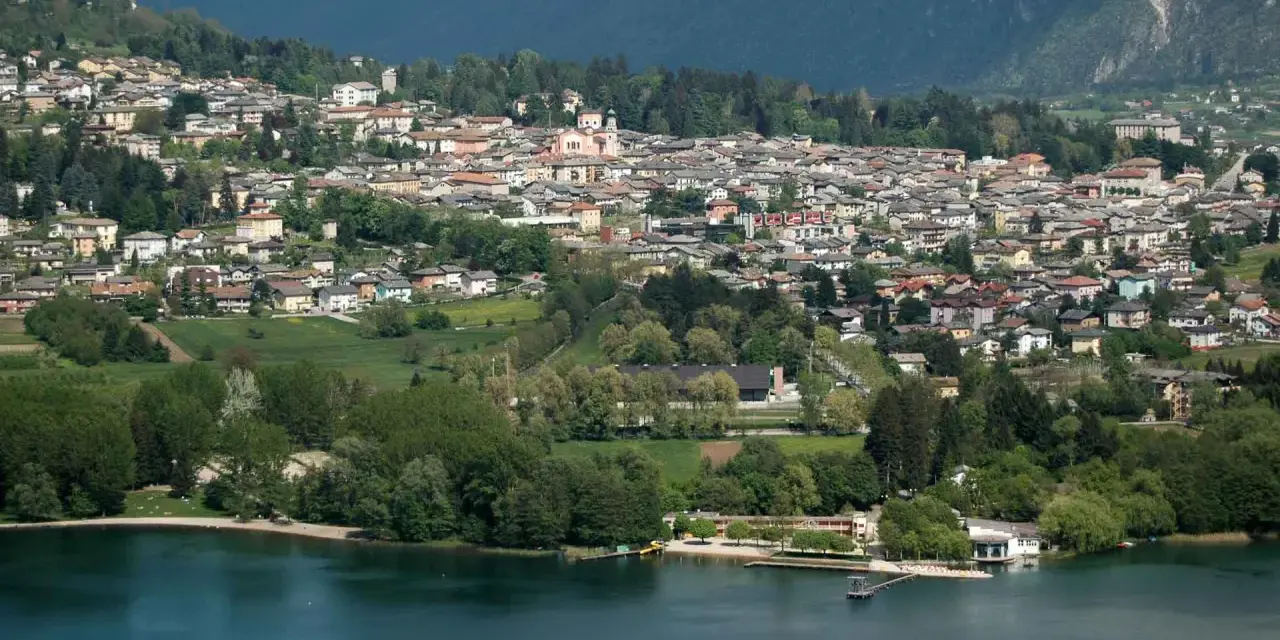 Vista panoramica del **lago di Levico** con il paese arroccato sulla collina. Alberi verdi e case colorate si specchiano nell'acqua tranquilla.