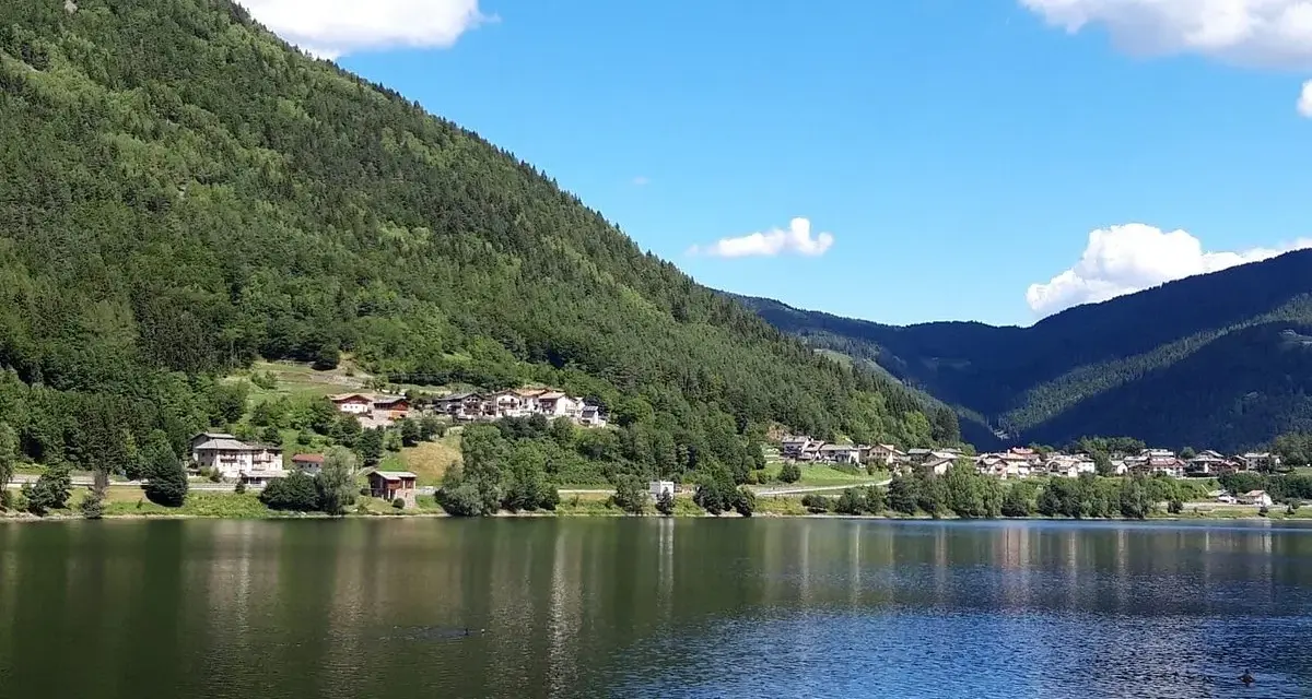 Il **lago di Serraia** riflette le montagne boscose e un villaggio alpino sotto un cielo azzurro con nuvole bianche.