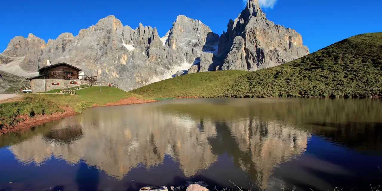 Riflessi maestosi delle cime nel lago di Val Venegia, con un rifugio alpino in primo piano.