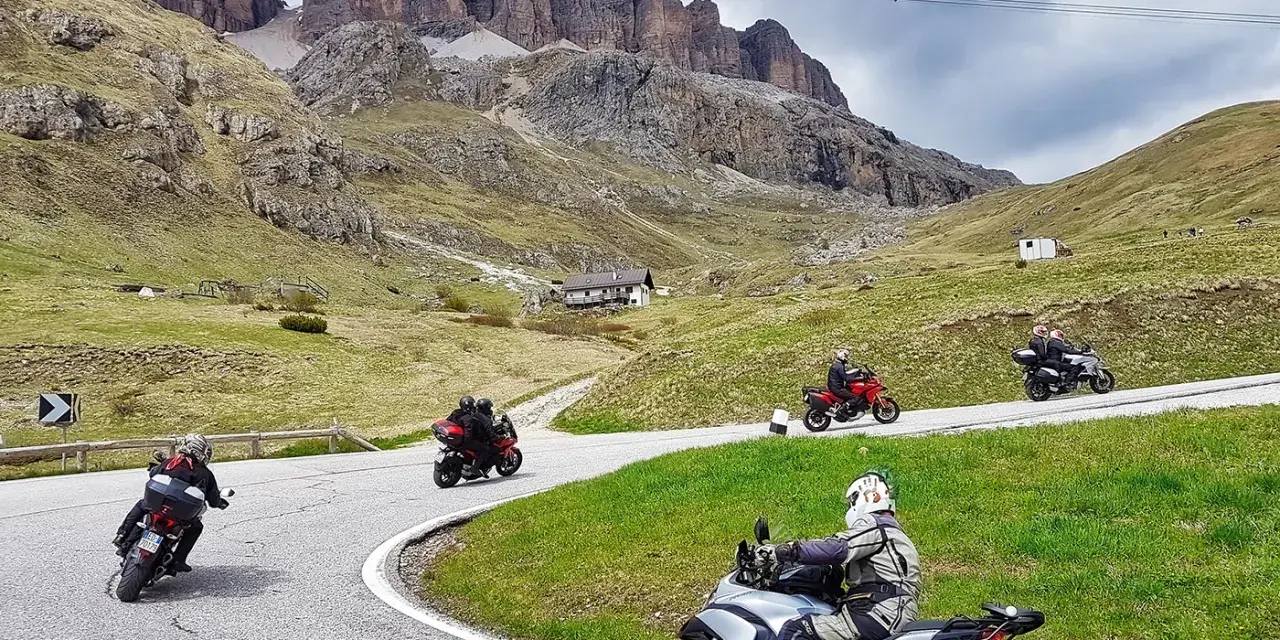 Motociclisti affrontano tornanti sui **Dolomiti passi**, con imponenti vette sullo sfondo.