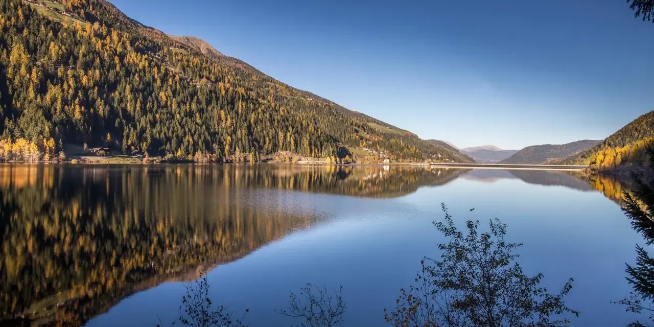 Autunno dorato sul **Val d'Ultimo lago**, con boschi che si specchiano nelle acque calme e un cielo azzurro intenso.