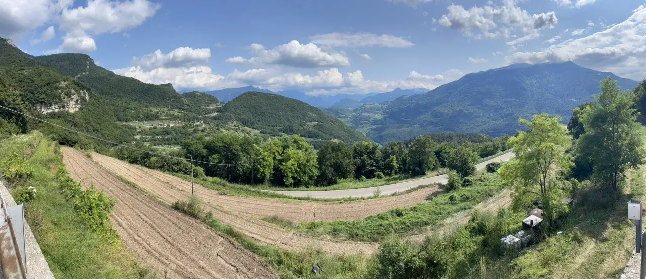 Panoramica del passo di Santa Barbara a Trento, con campi coltivati, alberi verdi e montagne sullo sfondo.