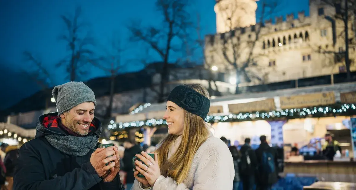 Coppia sorridente con tazze fumanti al mercatino di Natale di Trento inverno, con il castello illuminato sullo sfondo.