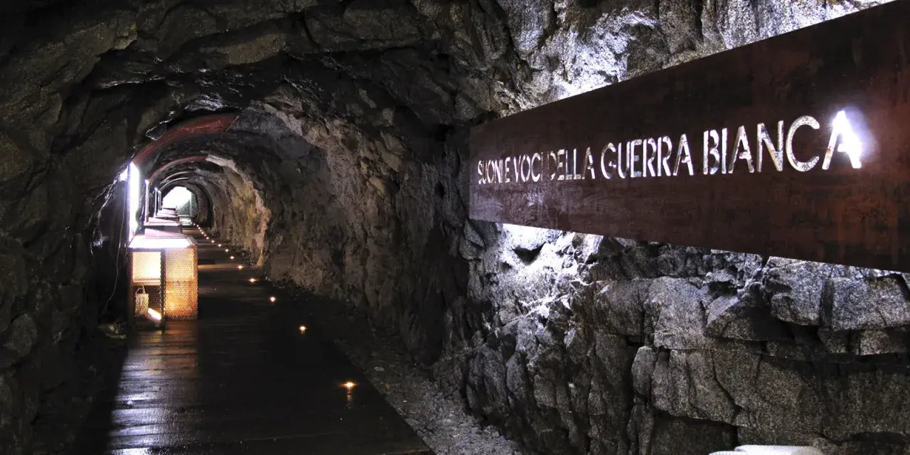 Un suggestivo tunnel roccioso illuminato, con un cartello che recita "Suoni voci della Guerra Bianca". Un percorso che sembra condurre alla galleria paradiso.